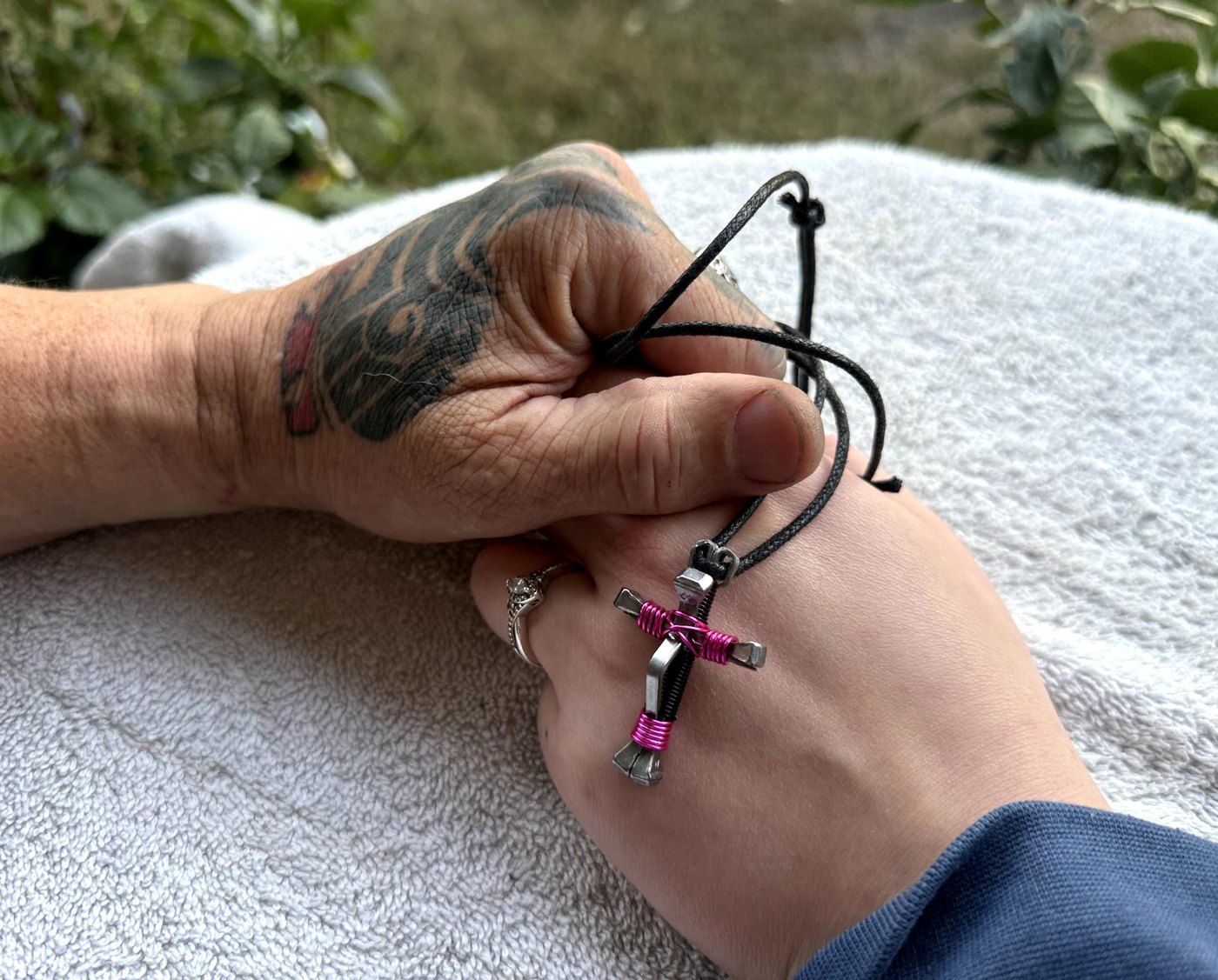 John's tattooed hand holding his daughter's hand with a pink horseshoe nail cross necklace